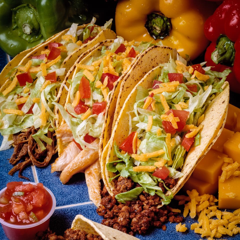 Three hard-shell tacos with various fillings, lettuce, cheese, and tomatoes, alongside salsa and bell peppers.