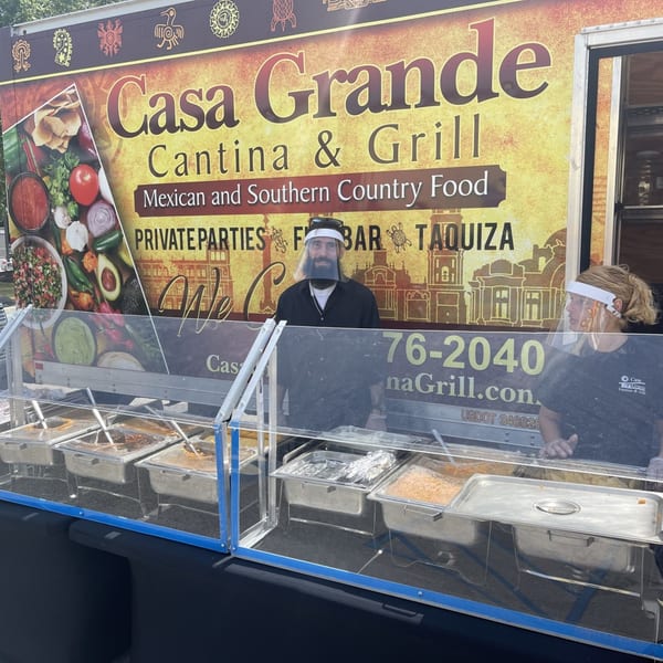Two Casa Grande Cantina & Grill staff members in face shields stand behind a buffet of Mexican food.
