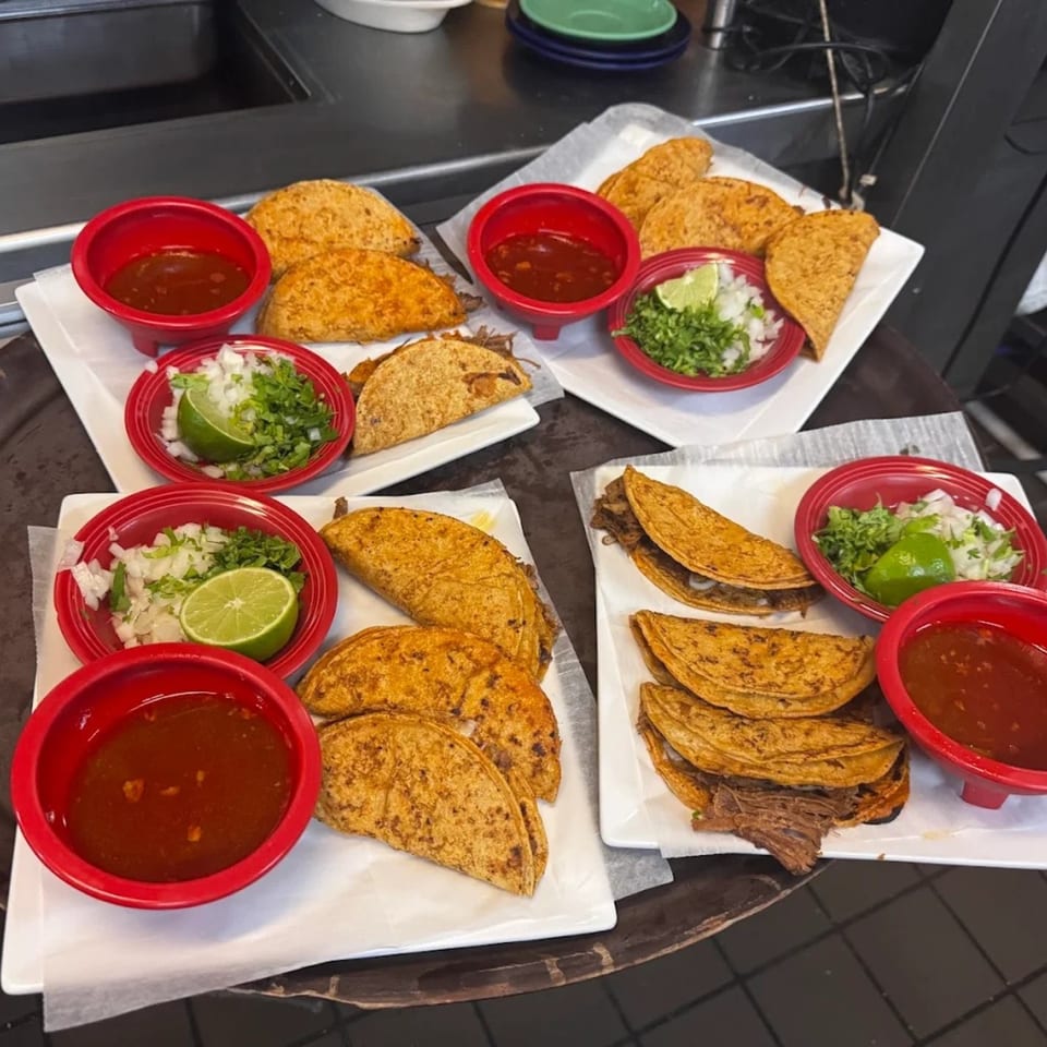 Multiple plates of golden-brown birria tacos with sides of red sauce, diced onions, cilantro, and lime.